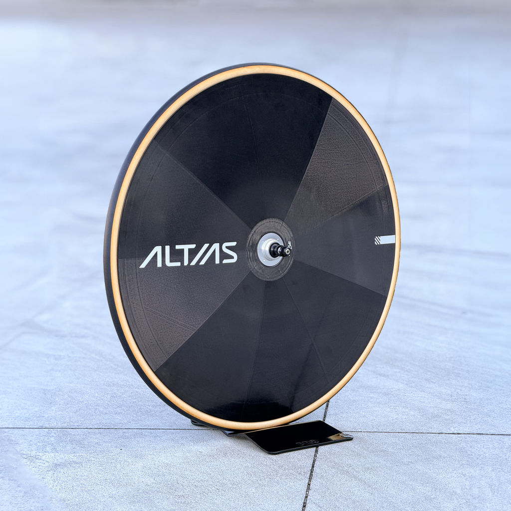 A black Altas disc wheek with a tan coloured tyre, displayed on a small black stand on a gray concrete floor.