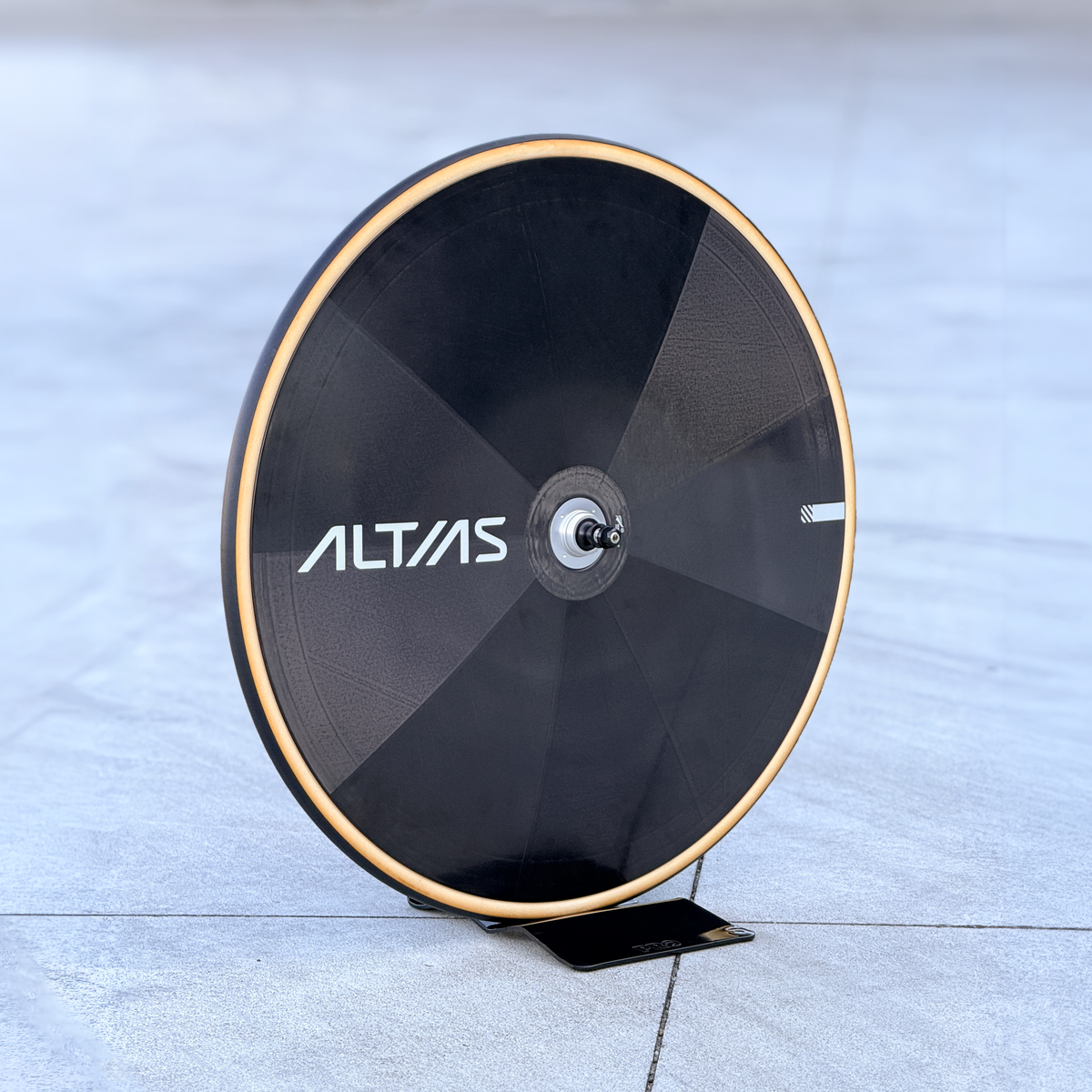 A black Altas disc wheek with a tan coloured tyre, displayed on a small black stand on a gray concrete floor.